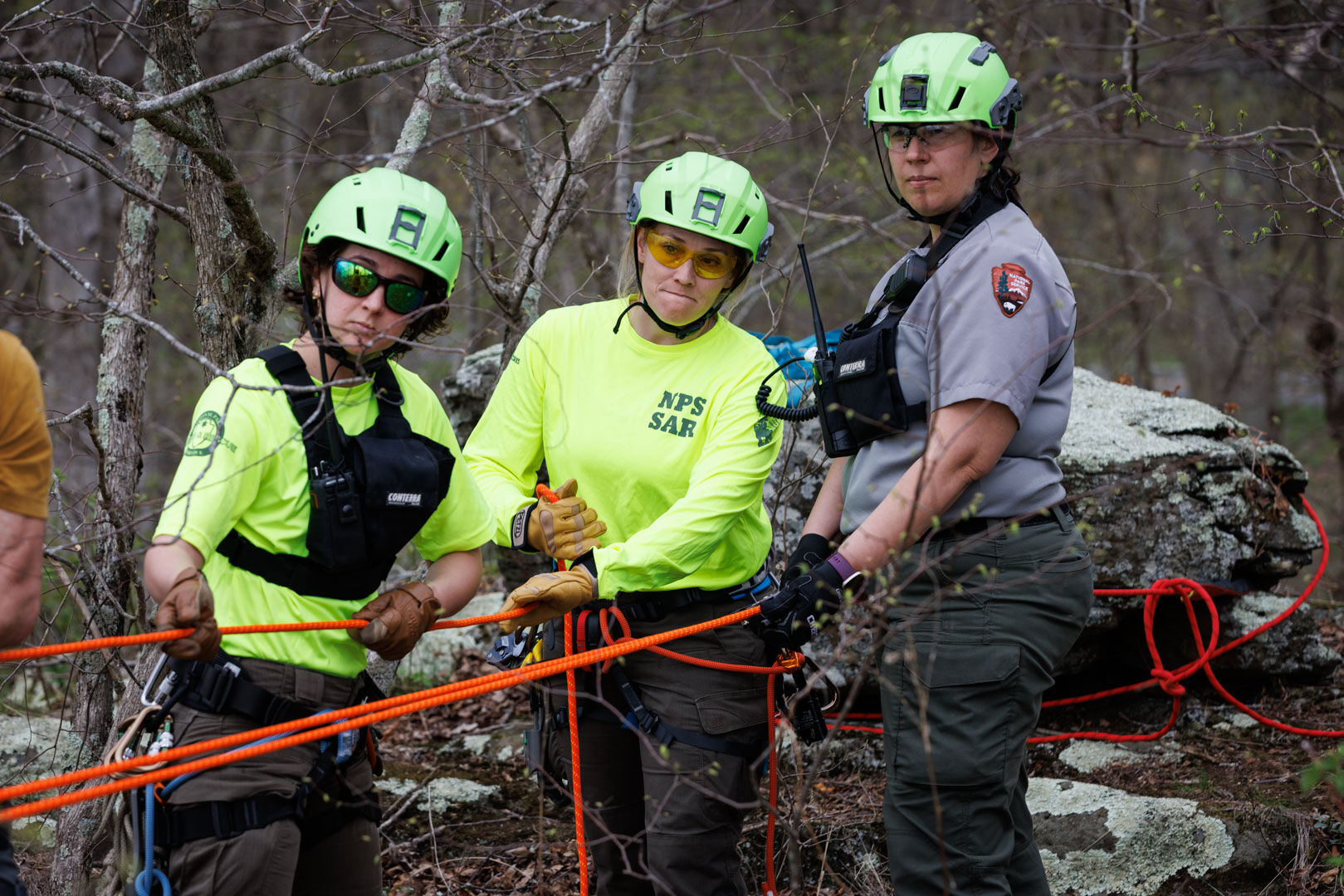 Ranger Avery Shirvani, Chief Ranger Cynthia Sirk-Fear and Ranger Kelly Good guiding the ropes tied to a boulder atop the cliff.