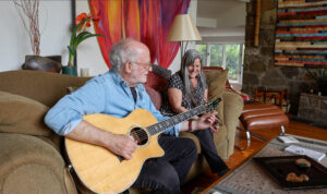 Paul Reisler tunes his guitar sitting next to wife Cheryl on a couch in their home