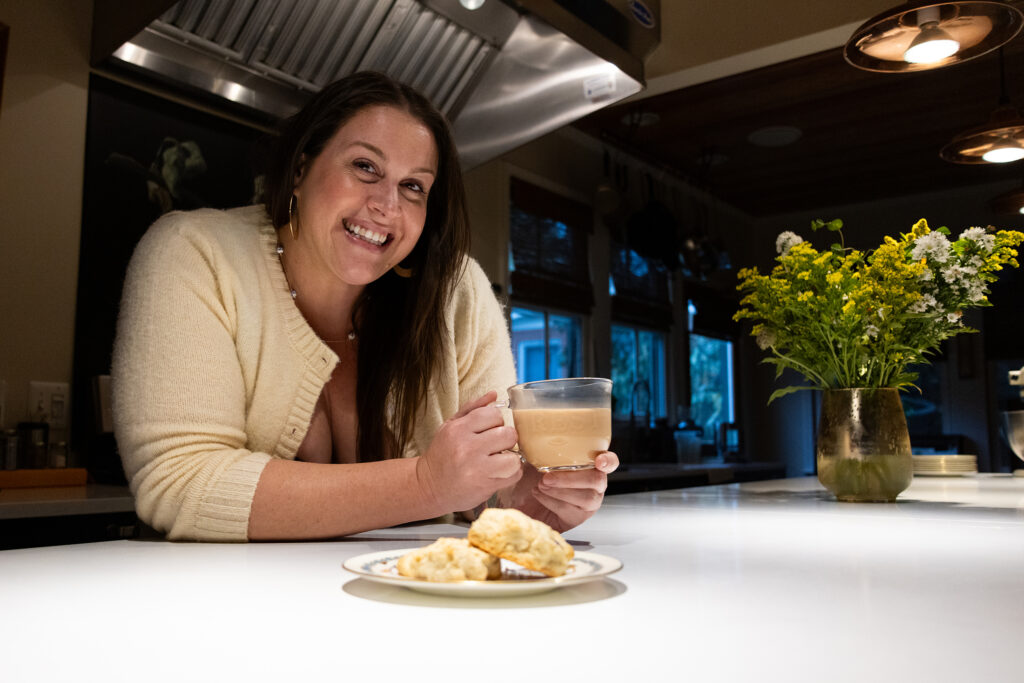 Huff standing, leaning on the kitchen island with a plate of scones in front of her and a cup of tea in her hands